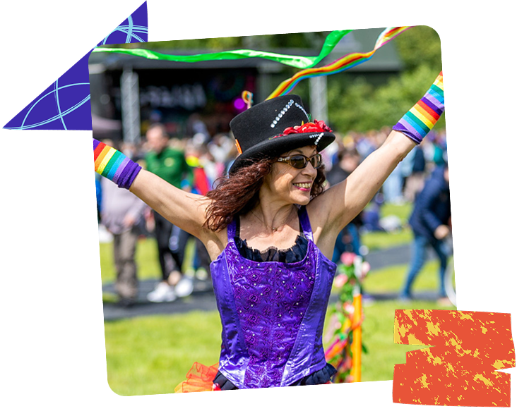 Woman wearing a colourful costume with rainbows and a hat, smiling with her arms open wide, as she walks through the crowd on a sunny day at Ablefest
